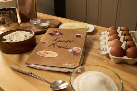 Cookbook surrounded by baking ingredients including flour, eggs, sugar and butter